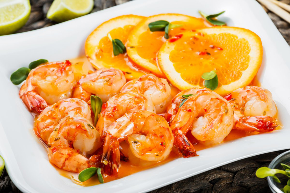 Frandon_26avril2021_AdobeStock_219448531_1200x_Honey Glazed Shrimp with orange slices, spices and sunflower sprouts on white plate on bamboo background. Food for Holidays.